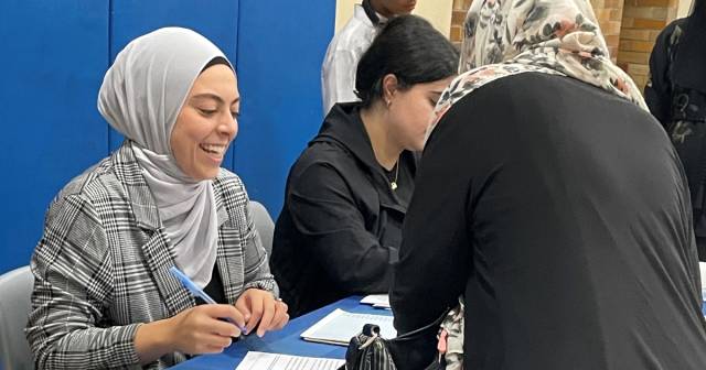 A woman in a grey hijab and gray jacket smiles and greets a woman 