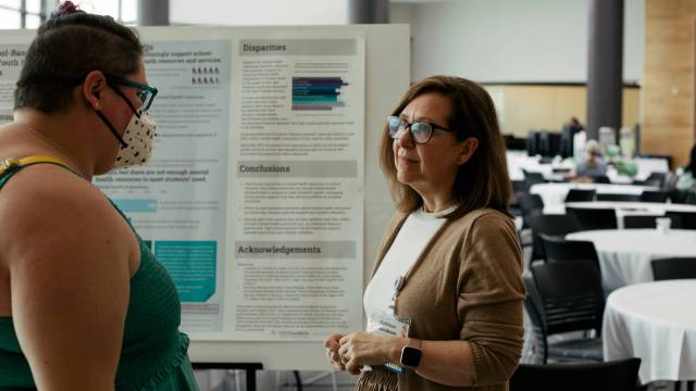 A woman stands in front of a scientific poster and presents it to a person in a green dress.