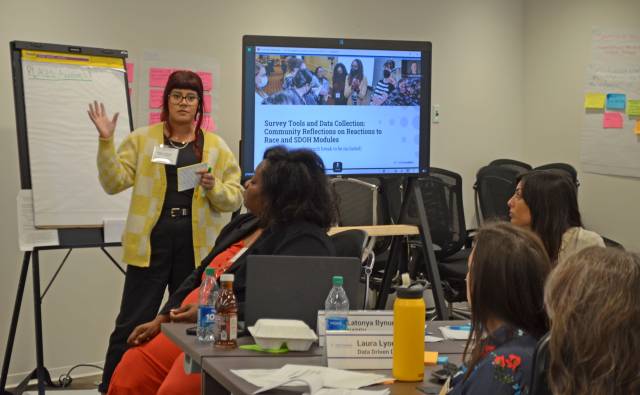 A woman in yellow stands in front of a large white board and talks to people sitting down.