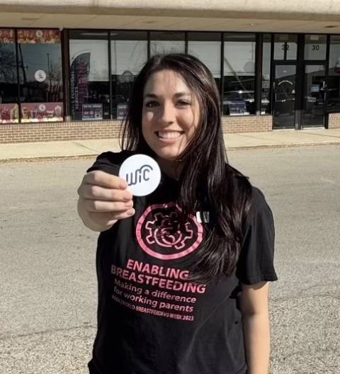 A smiling young brown haired young woman holds up a WIC sticker